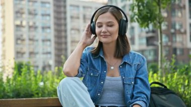 Kulaklıklı, müzik dinleyen, dışarıda bankta rahatça oturan mutlu genç bir kadın. Olumlu, neşeli, gözleri kapalı bir kadının portresi. Gençlik, teknoloji, yaşam tarzı, duygular