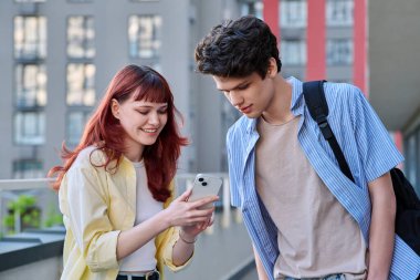 Arkadaşlar genç erkek ve kız üniversite öğrencileri birlikte konuşuyor, açık havada akıllı telefonlara bakıyorlar, şehrin modern binalarına bakıyorlar. Yaşam tarzı, iletişim, 19-20 yaş arası, şehir tarzı.
