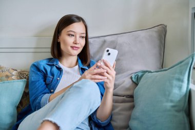 Evdeki koltukta oturan akıllı telefonu kullanan genç bir kadın. Kadın mesajlaşma görüntüsü, mobil uygulamalar teknoloji iş eğitimi için internet hizmetleri eğlence iletişim alışveriş yaşam tarzı