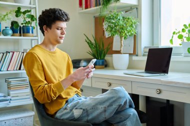 Genç adam akıllı telefon kullanıyor, evde oturuyor. Yakışıklı üniversite öğrencisi 19-20 yaşlarında, sarı saçlı, kıvırcık saçlı. Teknolojiler, mobil uygulamalar, iletişim eğlence kavramı