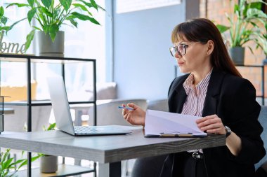 Orta yaşlı iş kadını laptopta görüntülü sohbet görüşmesi yapıyor ofis içi çalışma masasında oturuyor. Çevrimiçi iletişim teknolojisi işi, girişimcilik iş danışmanlığı