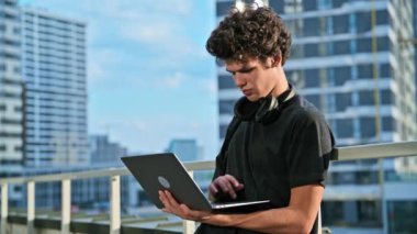 Dizüstü bilgisayarda yazan genç bir erkek, modern şehir gökdelenleri arka planı. Yakışıklı bir öğrenci, 19-20 yaşlarında, boş zamanlarında bilgisayar kullanıyor, okuyor, çalışıyor. Teknoloji, gençlik, eğitim, kentsel tarz kavramı