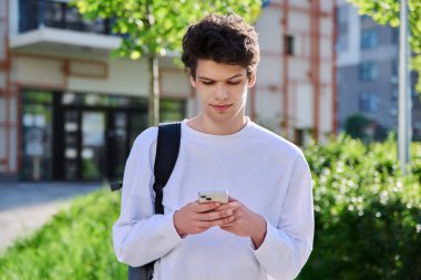 Açık havada akıllı telefon kullanan yakışıklı genç bir erkek. 19-20 yaşlarında sırt çantalı bir öğrenci. Eğitim, üniversite eğitimi, gençlik yaşam tarzı konsepti