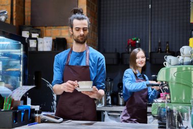 Önlüklü, kahve barının arkasındaki barın arkasında elinde fincanla gülümseyen yakışıklı bir erkek baristanın portresi. İş, yemek servisi, girişim, hizmet personeli, girişimcilik, küçük işletme konsepti