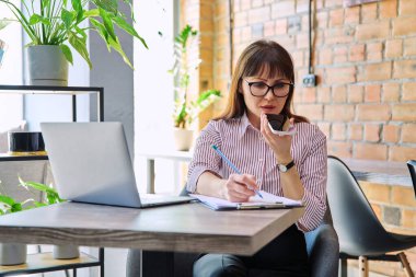 Orta yaşlı gülümseyen bir iş kadınının portresi dizüstü bilgisayarla çalışıyor, telefonla konuşuyor, ofis içinde masa başında oturuyor. İş, uzaktan kumanda, serbest çalışma, teknoloji, 40 'lı yaşlarda insanlar