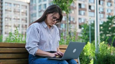 Orta yaşlı, gözlüklü, dizüstü bilgisayarda yazı yazan bir kadın. Kenar mahalle tarzı bir bankta oturuyor. Teknoloji, uzak ticaret, iş, eğlence, serbest çalışma, yaşam tarzı konsepti