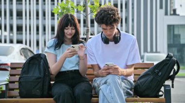 Ergen üniversite öğrencileri erkek ve kız birlikte konuşuyor, akıllı telefon kullanıyor, dışarıda oturuyor. 19-20 yaş arası, mobil uygulamalar teknolojiler, eğitim, yaşam tarzı, arkadaşlık kavramı.