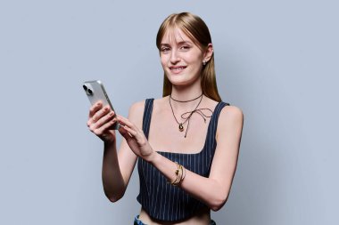 Gri stüdyo arka planında akıllı telefon kullanan güzel bir kadın. Havalı sarışın kadın cep telefonuyla kameraya bakıyor. Teknolojiler, mobil uygulamalar uygulamalar, hizmetler, gençlik kavramı