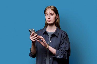 Genç ve güzel bir kadın mavi stüdyo arka planında akıllı telefon kullanıyor. Ciddi bir üniversite öğrencisi cep telefonuyla kameraya bakıyor. Teknolojiler mobil uygulama hizmetleri gençlik eğitimi kavramı