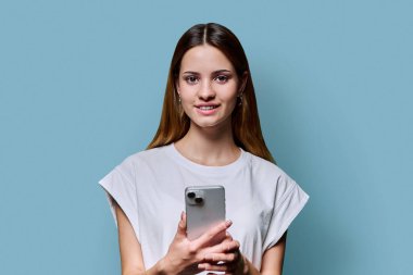 Genç ve güzel bir kadın mavi stüdyo arka planında akıllı telefon kullanıyor. Moda tebessüm eden kızıl saçlı kadın cep telefonuyla kameraya bakıyor. Teknolojiler mobil uygulama hizmetleri gençlik kavramı