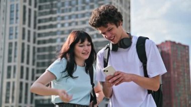 Arkadaşlar genç erkek ve kız üniversite öğrencileri birlikte konuşuyor, açık havada akıllı telefonlara bakıyorlar, şehrin modern binalarına bakıyorlar. Yaşam tarzı, iletişim, 19-20 yaş arası, şehir tarzı.