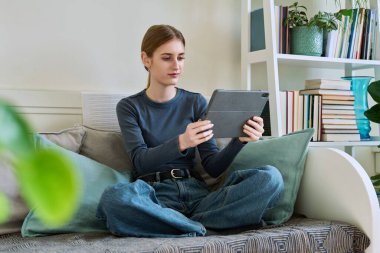 Genç bir bayan dijital tablet kullanıyor, evdeki kanepede oturuyor. Modern kablosuz teknolojiler, internet, çevrimiçi, uygulamalar, dijital aygıtlar