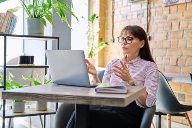 Ciddi şekilde konuşan orta yaşlı bir kadın laptopunu kullanarak görüntülü konuşma yapıyor, birlikte çalıştığı bir kafede oturuyor. İş, internet teknolojisi, başarı liderlik yönetimi yetkilendirmesi