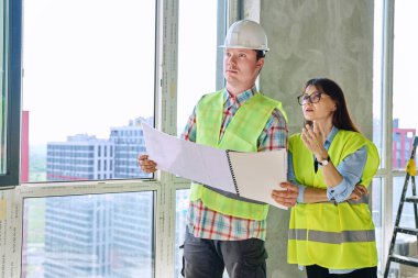 Koruyucu yelek kasklı iş arkadaşları yeni inşaat sahasında birlikte çalışarak plan belgelerini tartışıyorlar. Yönetim teftişi yenileme sektörü hizmet geliştirme işi