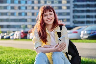 Sırt çantası açık havada akıllı telefon kullanan genç bir üniversite öğrencisi. Yüz piercingli kızıl saçlı modern hippi kız kameraya bakıyor. Moda, güzellik, eğitim, üniversite eğitimi, gençlik kavramı