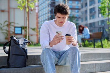 Rahatlamış, yakışıklı, akıllı telefonlu genç bir erkek, bir bardak kahve, merdivenlerde, dışarıda oturuyor. Sırt çantası, yaşam tarzı, gençlik konsepti olan 19-20 yaşlarında bir öğrenci.