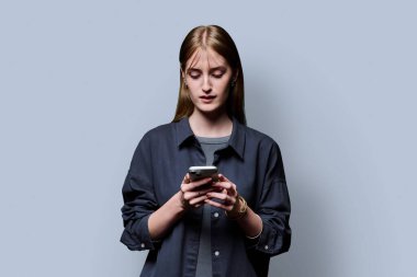 Gri stüdyo geçmişinde akıllı telefon kullanan genç ve güzel bir kadın. Ciddi bir üniversite öğrencisi cep telefonuyla kameraya bakıyor. Teknolojiler mobil uygulama hizmetleri gençlik eğitimi kavramı