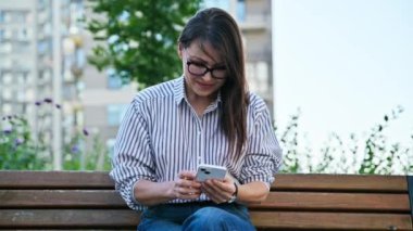 Açık havada akıllı telefon kullanan olgun, güzel bir kadın. Gülümseyen orta yaşlı bir kadın mesaj yazarken rahatça bankta oturuyor. Teknoloji mobil uygulamaları İnternet çevrimiçi yaşam tarzı
