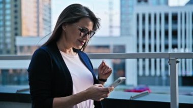 Akıllı telefon kullanan, modern şehir geçmişi olan olgun, ciddi bir iş kadını. Orta yaşlı başarılı gülümseyen kadın iş için mobil uygulama uygulaması kullanarak mesaj atıyor. Teknoloji İnternet işi kentsel