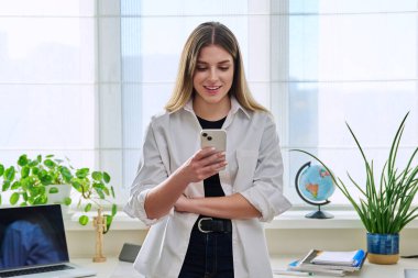 Pencerenin yanında duran akıllı telefonu kullanan genç bir kadın. Çalışmak için internet hizmetleri, iş eğitimi, iletişim alışverişi yaşam tarzı için kadın mesajları okuma, seyyar uygulama teknolojisi