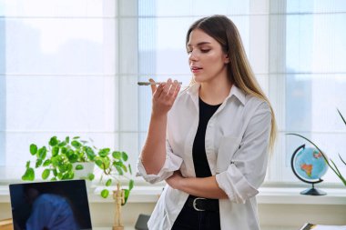 Genç bir kadın akıllı telefondan sesli mesaj kaydediyor. Evde, telefonun yanında konuşarak sesli mesaj gönderiyor. Teknoloji, iletişim, yaşam tarzı, gençlik kavramı