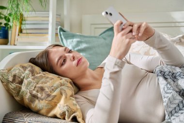 Mutlu gülümseyen, rahat ve rahat genç bir kadın evde kanepede uzanmış, akıllı telefonu eğlence iletişimi için kullanıyor. Gençlik, yaşam tarzı, dijital çevrimiçi teknoloji, cihazlar, internet konsepti