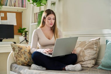 Evde çalışan genç bir kadın, dizüstü bilgisayar kullanarak kanepede oturuyor. Serbest meslek, eğitim, eğitim, uzaktan çalışma, boş zaman, internet teknolojisi uygulamaları konsepti