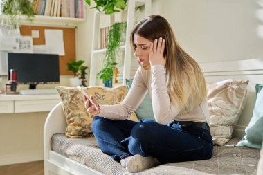 Evdeki kanepede oturan akıllı telefonu kullanan genç ve ciddi bir kadın. Telefon ekranına, mobil uygulamalara, internet teknolojisine bakan yorgun bir kadın. İş eğitimi için internet hizmetleri.