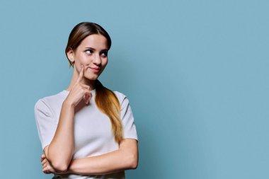 Genç, düşünceli, kızıl saçlı genç bir kadın mavi stüdyo arka planında mesaj için boş alana bakıyor. Gençler için ürün ve hizmetlerin reklamı
