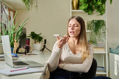 Genç bir kadın akıllı telefondan sesli mesaj kaydediyor. Evde, telefonun yanında konuşarak sesli mesaj gönderiyor. Teknoloji, iletişim, sesli mesaj, yaşam tarzı, gençlik kavramı