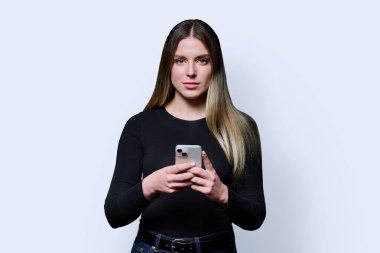 Cep telefonu tutan genç ve güzel bir kadın, beyaz stüdyo arka planındaki kameraya bakıyor. Gençlik, ağ interneti, iş, öğrenme, blog yazma, boş zaman, iletişim teknolojileri