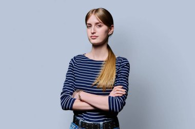 Gri stüdyo arka planında çapraz kollu güzel bir kızın portresi. Kendine güveni tam genç bayan sarışın kameraya bakıyor. Güzellik, yaşam tarzı, gençlik kavramı