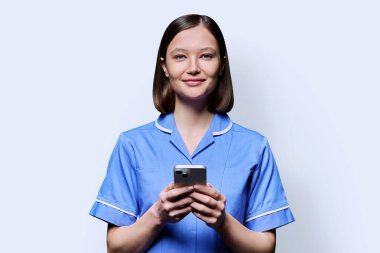 Akıllı telefon kullanarak gülümseyen genç bayan hemşire, beyaz stüdyo arka planındaki kameraya bakıyor. Mobil uygulamalar, tıbbi hizmetlerdeki teknolojiler, sağlık, profesyonel yardım, tıbbi bakım