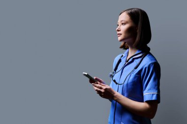 Akıllı telefon kullanan ciddi kadın hemşire, gri stüdyo arka planında profil görüntüsü, kopyalama alanı. Mobil uygulamalar, tıbbi hizmetlerdeki teknolojiler, sağlık, profesyonel yardım, tıbbi bakım
