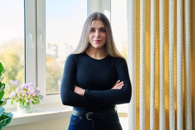 Kollarını kavuşturup pencerenin yanındaki kameraya bakan kendine güvenen güzel bir kadının portresi. Gençlik, güzellik, yaşam tarzı konsepti