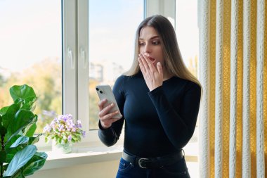 Pencerenin yanında duran akıllı telefonu kullanan genç, şaşkın bir kadın. Kadın mesajlaşma görüntüsü, mobil uygulama teknolojisi iş eğitimi için internet hizmeti boş iletişim alışverişi