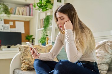 Evdeki kanepede oturan akıllı telefonu kullanan genç ve ciddi bir kadın. Telefon ekranına, mobil uygulamalara, internet teknolojisine bakan yorgun bir kadın. İş eğitimi için internet hizmetleri.