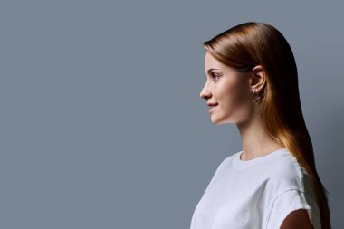 Kızıl saçlı güzel bir kızın profil portresi, gri stüdyo arka planı. Reklam boşluğuna bakan genç bir kadın. Moda güzellik eğitimi için ürünler ve hizmetler