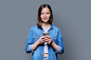 Genç mutlu kadın gri arka planda akıllı telefon kullanıyor. Gülümseyen 20 'li yaşlarda bir kadın kameraya mesaj atıyor. Mobil İnternet uygulamaları, iş eğitimi, eğitim, iletişim, sağlık hizmetleri alışveriş uygulamaları teknolojileri