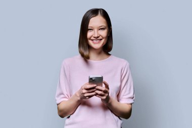 Genç mutlu kadın gri arka planda akıllı telefon kullanıyor. Gülümseyen 20 'li yaşlarda bir kadın kameraya mesaj atıyor. Mobil İnternet uygulamaları, iş eğitimi, eğitim, iletişim, sağlık hizmetleri alışveriş uygulamaları teknolojileri