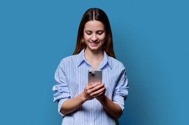 Genç ve güzel bir kadın mavi stüdyo arka planında akıllı telefon kullanıyor. Moda tebessümü olan kızıl saçlı bir kadın cep telefonuna bakıyor. Teknolojiler mobil uygulama hizmetleri gençlik kavramı