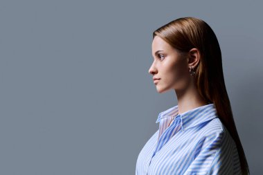 Kızıl saçlı güzel bir kızın profil portresi. Gri stüdyo arka planı. Reklam boşluğuna bakan ciddi bir genç kadın. Moda güzellik eğitimi için ürün sunar.