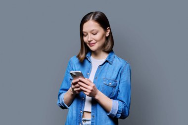 Genç mutlu kadın gri arka planda akıllı telefon kullanıyor. Gülümseyen 20 'li yaşlarda bir kadın telefona bakıyor, mesaj atıyor. Mobil İnternet uygulamaları, iş eğitimi, eğitim, iletişim, sağlık hizmetleri alışveriş uygulamaları teknolojileri