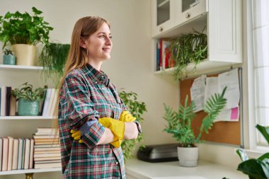 Ev temizliği için koruyucu lastik eldiven giyen, kollarını kavuşturup pencereye bakan genç bir kadının portresi. Temizlik, rutin, yaşam tarzı, gençlik kavramı