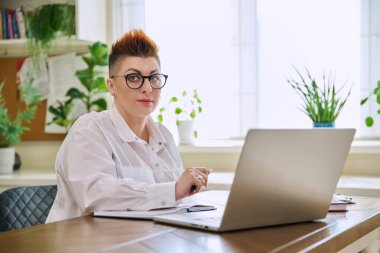 Evde bilgisayarla çalışan olgun bir iş kadınının portresi. Orta yaşlı gülümseyen bir kadın ofis masasında oturmuş kameraya bakıyor. Uzak iş çalışmaları, kariyer, yönetim, pazarlama