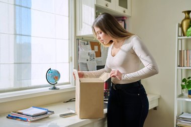 Evde alışveriş yapan genç bir bayan, içinde online alışverişler olan karton kutuları açıyor. Mutlu yirmili yaşlarda kutuları açan kadın tüketiciler. Postayla teslimat, çevrimiçi mağaza, e-alışveriş konsepti