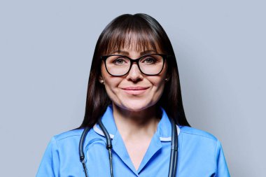 Gri stüdyo arka planında steteskobu olan mavi elbiseli, gülümseyen orta yaşlı kadın hemşire portresi. Tıbbi hizmetler, nhs, sağlık, mesleki yardım, tıbbi bakım kavramı