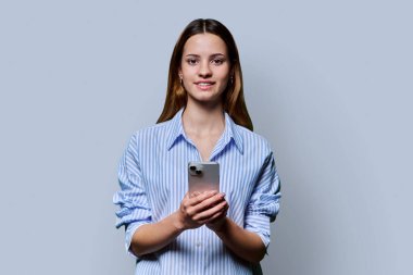 Gri stüdyo geçmişinde akıllı telefon kullanan genç ve güzel bir kadın. Moda tebessüm eden kızıl saçlı kadın cep telefonuyla kameraya bakıyor. Teknolojiler mobil uygulama hizmetleri gençlik kavramı