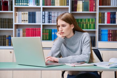 Eğitim kütüphanesinde dizüstü bilgisayar kullanarak okuyan ciddi bir genç kız öğrenci. Eğitim, ergenlik, lise, üniversite, öğrenim, bilgi kavramı