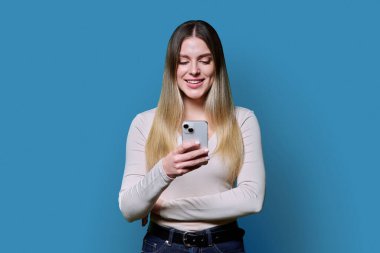 Cep telefonu kullanan genç ve güzel bir kadın mavi stüdyo geçmişindeki akıllı telefona bakıyor. Gençlik, ağ interneti, iş, öğrenme, blog yazma, boş zaman, iletişim teknolojileri